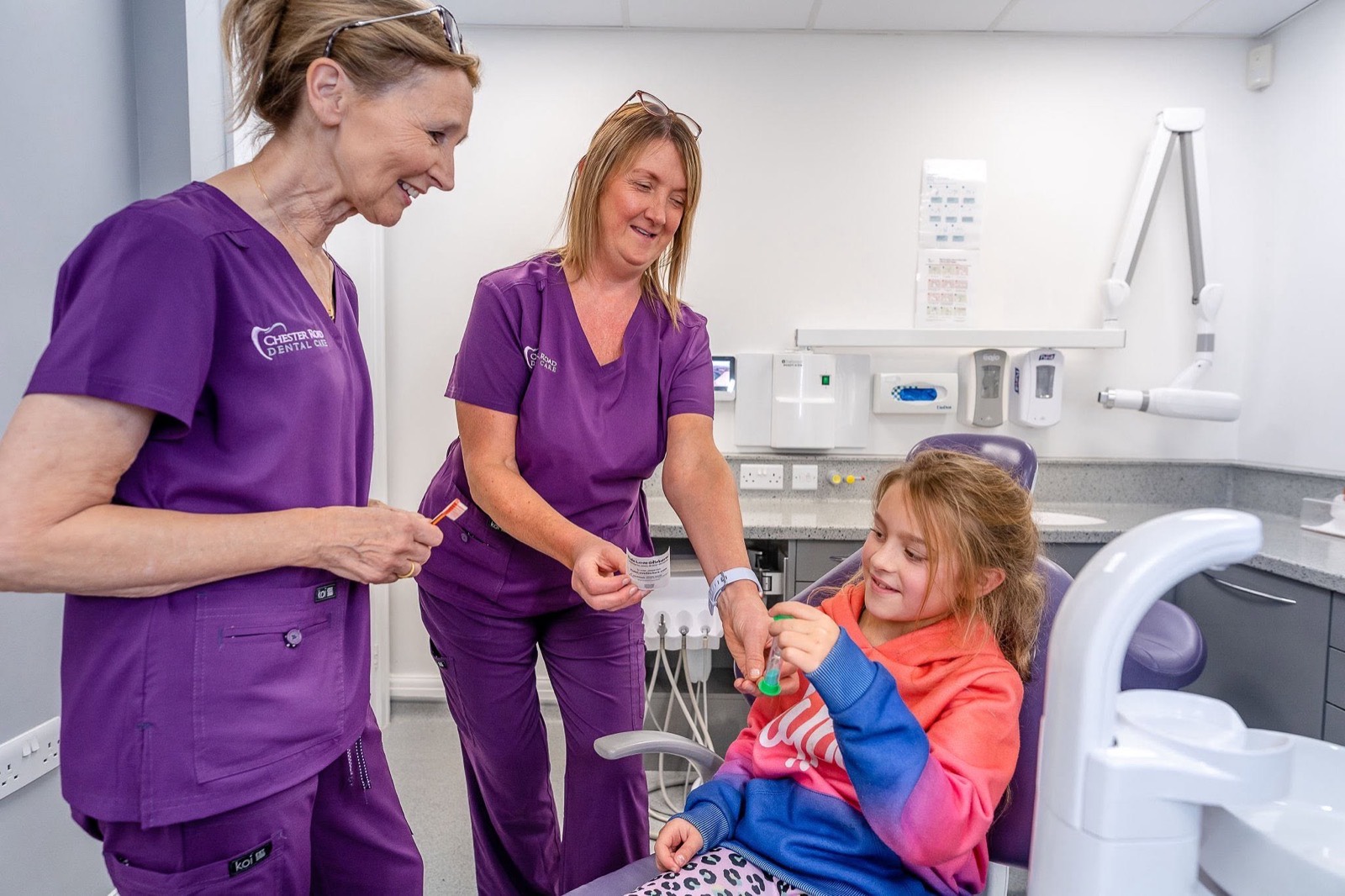 Two dental nurses in purple scrubs chatting with a young patient in the treatment chair